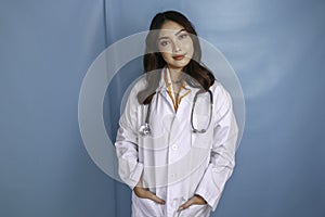 Confident relaxed female Asian doctor standing with hands in the pockets of her lab coat smiling quietly at the camera over blue