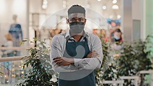 Confident restaurant worker in medical mask standing indoors barman seller waiter in apron turns to camera young