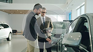 Confident sales manager talking to male customer in car dealership showing auto. Focus on vehicle mirror