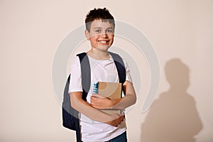 Confident schoolboy dressed in white mockup t-shirt and blue jeans, carrying backpack, holds copybooks, smiles at camera