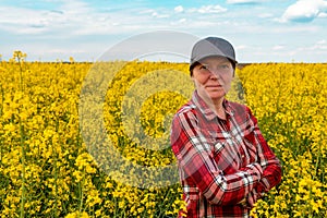 Confident and self-assured farm worker wearing red plaid shirt and trucker`s hat standing in cultivated rapeseed field in bloom