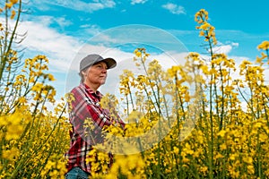 Confident and self-assured farm worker wearing red plaid shirt and trucker`s hat standing in cultivated rapeseed field in bloom