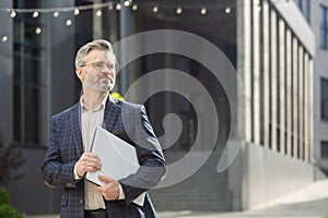 Confident senior businessman with laptop outside corporate building