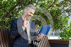 Confident senior businessman smiling and waving while using tablet outside office