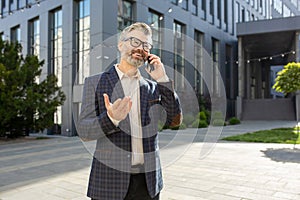 Confident senior businessman in suit communicating on smartphone outside modern office