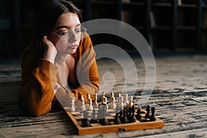 Confident sexy young woman in elegant eyeglasses thinking about chess move lying on wooden floor in dark room, looking