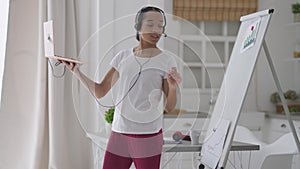 Confident slim beautiful woman in headphones pointing at business graphs on whiteboard talking at laptop web camera in