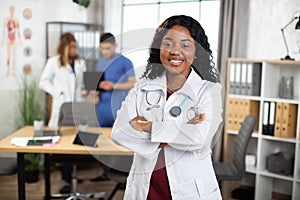 Afro-American doctor posing with arms crossed and diverse medical team working on the background