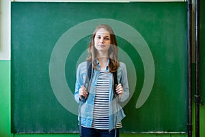 Confident smiling female high school student standing in front of chalkboard in classroom, wearing backpack, looking at camera.