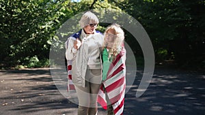 Confident smiling grandmother and granddaughter in American flag looking at camera standing in sunshine. Portrait of