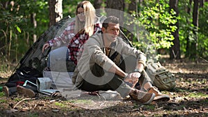 Confident smiling man sitting in sunshine in forest as woman getting out camp tent in slow motion. Wide shot portrait of