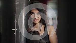 Confident smiling Middle Eastern sportswoman looking at camera kicking punching bag. Portrait of beautiful female boxer