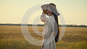 Confident smiling young european woman in light dress puts on hat and smiling. Enjoying nature at sunset near