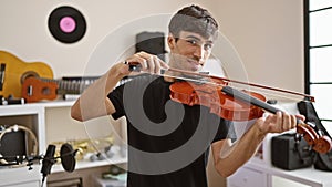 Confident and smiling young hispanic man artist playing violin happily in an indoor music studio, a symphony of classical
