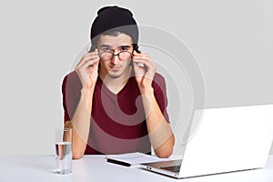 Confident student sits at workplace, looks attentively through round transparent glasses, dressed in stylish clothes, makes notes
