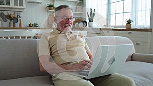 Confident stylish happy middle aged senior man using laptop at home. Stylish older mature 60s beard grandfather sitting