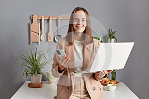 Confident successful woman freelancer with brown hair wearing beige jacket standing in home kitchen interior working online