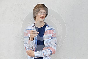 Confident teenage boy with stylish hairdo wearing casual shirt pointing with his finger at camera while showing something. Stylish