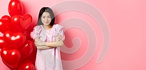 Confident teenage girl cross arms on chest and smile, celebrating valentines day in cute dress with red heart balloons