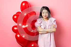Confident teenage girl cross arms on chest and smile, celebrating valentines day in cute dress with red heart balloons