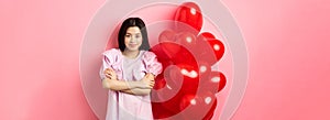 Confident teenage girl cross arms on chest and smile, celebrating valentines day in cute dress with red heart balloons