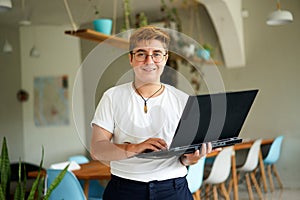 Confident transgender office worker with laptop in modern workspace. FTM professional in casual attire stands, ready for photo