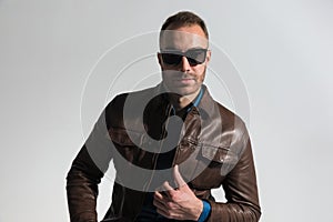 Confident unshaved guy with sunglasses fixing brown leather jacket