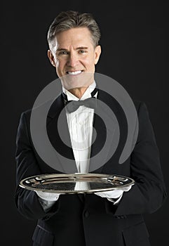 Confident Waiter In Tuxedo With Tray Standing Against Black Back