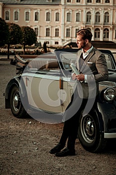 Confident wealthy young man with newspaper near classic convertible