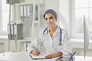 Confident woman doctor pediatrician writes in a clipboard sitting at a table in a white office of the hospital