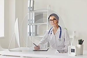 Confident woman doctor pediatrician writes in a clipboard sitting at a table in a white office of the hospital