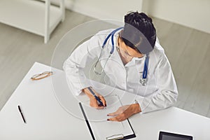 Confident woman doctor pediatrician writes in a clipboard sitting at a table in a white office of the hospital