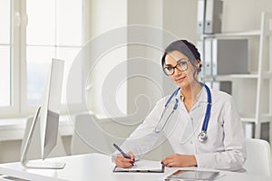 Confident woman doctor pediatrician writes in a clipboard sitting at a table in a white office of the hospital