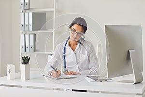 Confident woman doctor pediatrician writes in a clipboard sitting at a table in a white office of the hospital