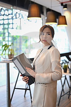Confident woman entrepreneur holding file folder and looking away while standing in modern workplace.