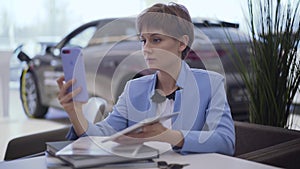 Confident woman with short hair in classic blue formal suit sits at the table talking by cell phone using front camera