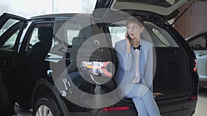 Confident woman with short hair in classic blue formal suit sitting in open car trunk talking by cell phone. Girl