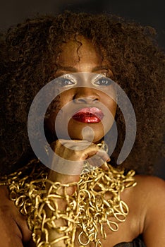 Confident young African woman with gold earthy makeup and necklace, laying hands on her chin, straight looking