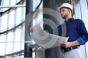 Confident young architect in navy shirt and hardhat holding a blueprint and looking at it