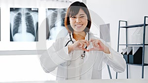 Confident young Asia female doctor in white medical uniform making a heart gesture with her fingers and smiling while video