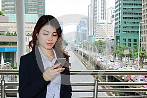 Confident young Asian business woman looking mobile smart phone in her hands at urban building city background.