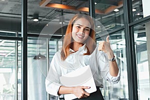 Confident young Asian business woman shows thumbs up hands sign in office