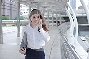 Confident young Asian business woman talking on phone while walking on walkway of modern city.