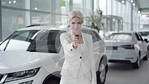 Confident young blond woman showing car keys at camera and smiling. Businesswoman in white suit buying vehicle in