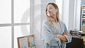 Confident young blonde woman worker standing with arms crossed, throwing a successful smile, while looking through the office