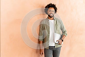 Confident young business man carrying laptop standing over beige wall, smiling proud male office employee in casual
