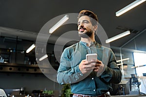 Confident young business man using mobile phone standing in office looking away.