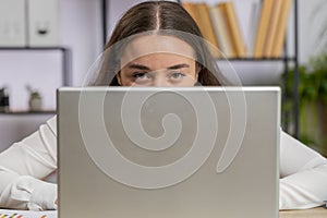 Confident young business woman hiding behind laptop computer, looking at camera, spying, peeping