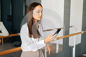 Confident young business woman in stylish clothes using browsing mobile phone standing in room with mirror, looking on