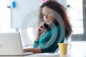 Confident young business woman talking on mobile phone while working with her laptop in the office